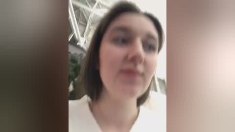 Media: Blurry video of a Caucasian woman with shoulder-length brown hair, wearing a white top, standing in a modern indoor setting with metallic structures and green plants in the background.