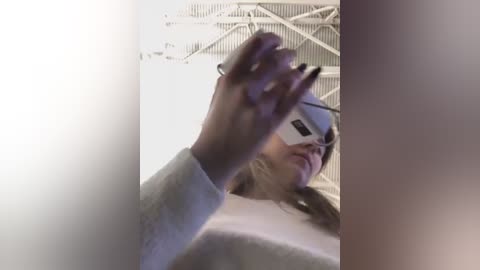 Media: Video of a woman with light skin and straight brown hair, wearing a white virtual reality headset, a grey sweater, and black nail polish, captured from a low-angle perspective. The background features an industrial white ceiling with exposed beams.