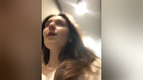 Media: A video shows a close-up of a young woman with long brown hair, wearing a white top, looking up with a slightly open mouth, in a dimly lit room.