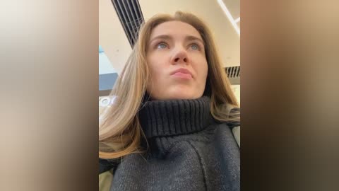 Media: Video of a young woman with fair skin, blue eyes, and blonde hair, wearing a dark sweater, looking upward with a contemplative expression, inside an airplane cabin with beige walls and black blinds.