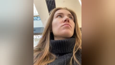 Media: A video of a Caucasian woman with long, light brown hair, wearing a dark, ribbed turtleneck sweater, gazing upward with a contemplative expression. The background features a modern, white-walled room with a partially visible sign and large windows.