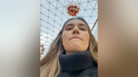 Media: Video of a Caucasian woman with straight, light brown hair, wearing a dark navy turtleneck sweater, standing in a modern, geometrically patterned glass atrium with a hanging, red and gold ornament.