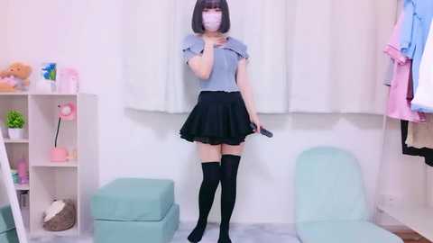 Media: Video of an Asian woman with straight black hair, wearing a blue blouse, black skirt, and thigh-high socks, standing in a pastel-colored room with white curtains, pink plant, and mint green chair.