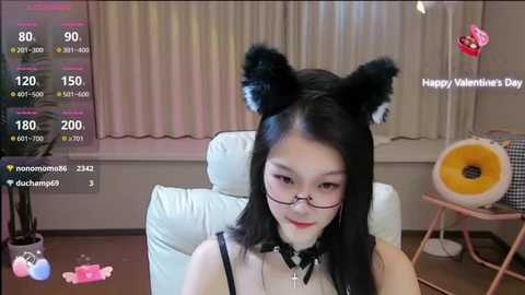 Media: Video of an Asian woman with black cat ears, glasses, and a black choker, sitting on a white chair, wearing a black top, in a dimly-lit room with a pink heart-shaped pillow and a pink table.