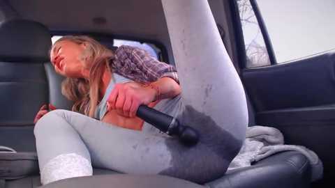 Media: Video of a blonde woman in a car, wearing a plaid shirt, grey leggings, and white socks, using a black vibrator on her crotch.