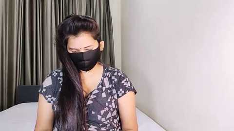 Media: Video of an East Asian woman with long black hair, wearing a black face mask and a patterned grey shirt, seated in a minimalist room with beige walls and grey curtains.