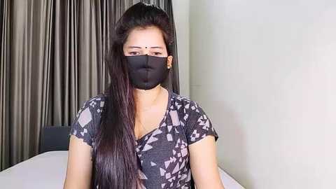 Media: Video of a woman with long black hair, wearing a black face mask, black patterned top, and sitting indoors against a beige wall and dark curtains.