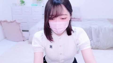 Media: Video of an Asian woman with straight, shoulder-length black hair and pale skin, wearing a white blouse and pink mask, in a soft, pastel-toned bedroom with white furniture and bedding.