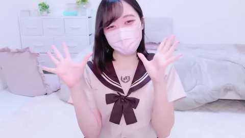 Media: A young Asian woman with straight black hair and pale skin, wearing a pink surgical mask, poses playfully in a white bedroom. She has a pink sailor school uniform with a large bow.