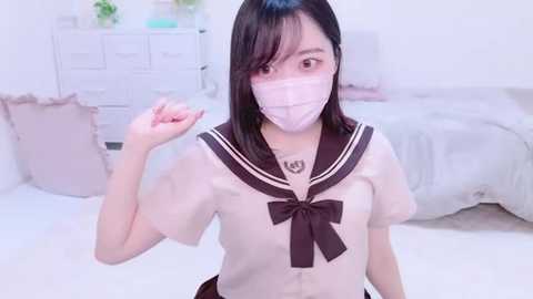 Media: Video of a young East Asian woman with short black hair, wearing a pink surgical mask, navy school uniform, and brown bow. She's indoors in a bright, white bedroom with a bed, dresser, and plants.