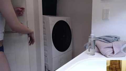 Media: Video of a small, modern laundry room with a person wearing a white tank top and blue shorts opening a white cabinet door. A white dryer and folded laundry are visible.