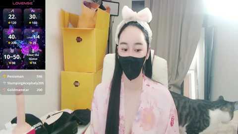 Media: Video of an East Asian woman in a pastel pink robe with a cat headband, wearing a black face mask, seated on a white chair in a cozy, dimly lit room with a cat on a blanket.