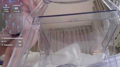 Media: A video of a transparent baby crib with a white blanket and a woman in a sheer, patterned dress, standing beside it, taken indoors with a soft, ambient lighting.