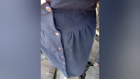 Media: Video of a person's midsection wearing a navy blue, pleated skirt with a fitted waistband, brass buttons, and a subtle texture, standing outdoors on a stone-paved path.