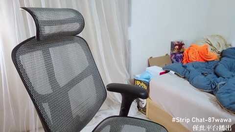 Media: Video of a messy bedroom with a black mesh office chair, a disheveled bed with clothes and a box, and white curtains in the background.