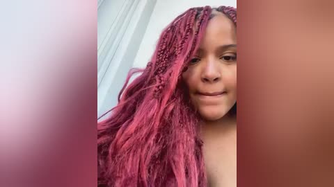 Media: Video of a Black woman with long, wavy, vibrant red braids partially covering her face. She has a neutral expression. The background is blurred with soft pink and peach tones.