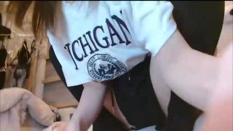 Media: Video of a young woman in a white tee with \"CHIGAN\" printed in black, bending over, exposing her midriff. Background shows a messy room with clothes scattered on the floor and a wooden staircase.