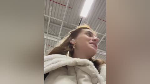 Media: A video of a woman with long blonde hair wearing a white coat and a furry collar, standing in a dimly lit, industrial warehouse with a corrugated metal ceiling and exposed pipes.