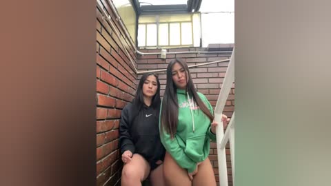 Media: Video of two Asian women with long black hair, sitting on a brick wall in a stairwell. One wears a black hoodie, the other a green hoodie, both exposing their thighs.