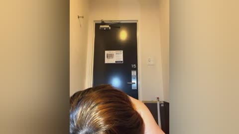 Media: A video shows a hotel hallway with beige walls and a black door with a \"15\" number plate. A person with brown hair, seen from the back, is standing near the door.