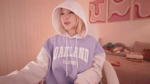 Media: Video of a blonde woman wearing a light blue hoodie with white sleeves, hood up, in a cozy living room with beige walls and abstract pink art.