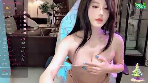 Media: Video of an East Asian woman with long black hair, fair skin, and small breasts, topless, covering her breasts with her hands. She's indoors, in a modern room with a Christmas tree and a kitchen in the background.