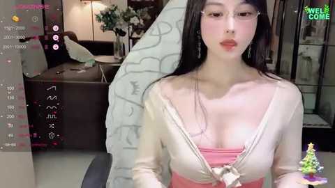 Media: Video of an Asian woman with long black hair, fair skin, wearing glasses, a pink top, and a white cardigan, standing in a modern room with a large mirror, digital interface, and a colorful Christmas tree.