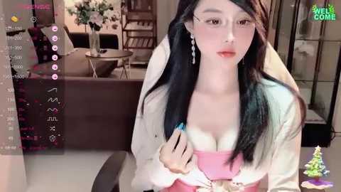 Media: A video of an East Asian woman with long black hair, fair skin, and a slender build, wearing a pink strapless top, white cardigan, and dangling earrings, sitting in a modern living room with a beige sofa, floral arrangement, and bookshelves.