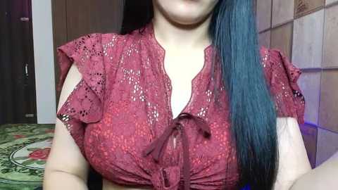 Media: A video of a woman with fair skin and long, straight, dark blue hair. She wears a red lace crop top with a deep V-neck, revealing ample cleavage. The background features a tiled wall and a carpet with intricate patterns.