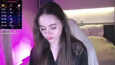 Media: Video of a young woman with long brown hair, wearing a black lace top, sitting on a white couch in a dimly lit room, with a purple light.