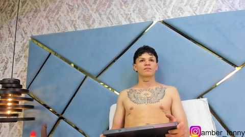 Media: Video of a shirtless, tattooed young man with short dark hair, holding a tablet, in a modern, geometric-patterned room with a blue, diamond-shaped headboard and a hanging pendant lamp.