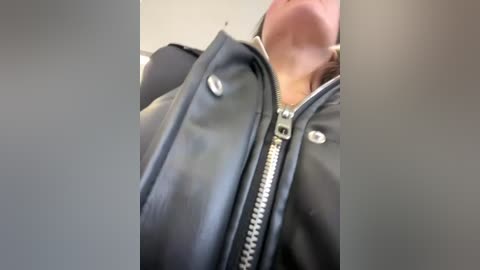 Media: A close-up video of a person wearing a black leather jacket with a silver zipper, partially open to reveal a light-colored shirt underneath. The background is blurred, focusing attention on the jacket's texture and the person's upper body.