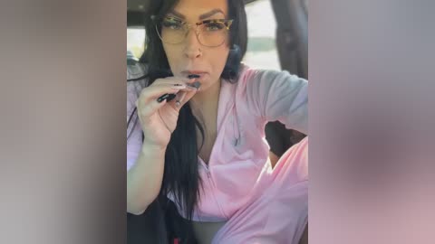 Media: A video of a woman with long black hair, wearing glasses, and a light pink hospital gown, sitting in a car. She has a serious expression and is holding a cigarette to her mouth.