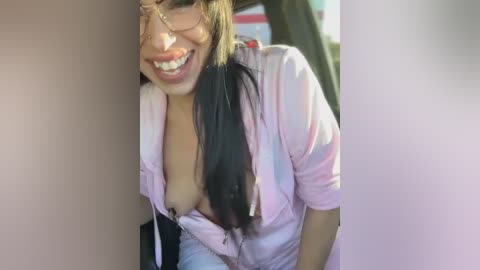 Media: A video of a smiling Asian woman with long black hair, wearing a pink shirt unbuttoned to reveal her large breasts. She is leaning against a beige wall, indoors.