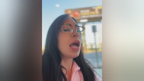 Media: A video of a young woman with long black hair, wearing glasses, pink shirt, and open mouth, standing in front of a blurred fast-food restaurant sign.