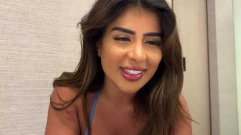 Media: Video of a smiling Latina woman with long brown hair, wearing a blue top, against a beige textured wall. She has dark eyebrows, dark eyes, and a light skin tone.