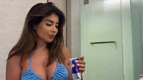 Media: A video of a Latina woman with long, dark hair, wearing a blue, ruffled bikini top, spraying a can of aerosol. The background features a beige wall and a frosted glass door.