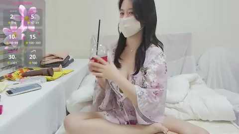 Media: Video of an East Asian woman with long black hair, wearing a pink floral robe, white mask, and a red drink, sitting cross-legged on a white bed, surrounded by cluttered bedside table items.