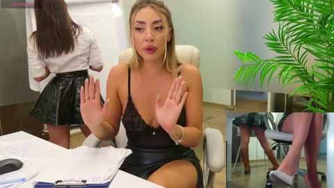 Media: Video of a blonde woman in a black lace dress, holding her hands up in a meeting room, with another woman in a white shirt and black skirt, and a green plant in the background.