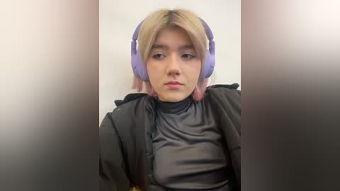 Media: Video of a young Asian woman with light skin, platinum blonde hair, and purple headphones, wearing a black high-necked top. She has a neutral expression against a blurred, gradient background.