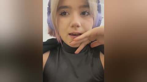 Media: A close-up video of a young woman with fair skin, platinum blonde hair, and purple headphones, wearing a black sleeveless top, with a blurred background.