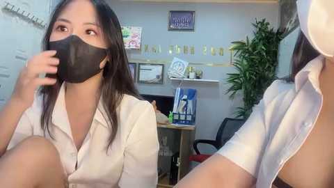 Media: Video of two Asian women in an office, one with a black face mask, light skin, and long black hair, and the other with a white shirt, partially unbuttoned, revealing cleavage. Background shows a desk with papers, plants, and framed certificates.