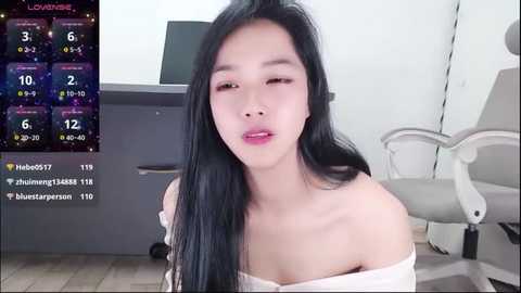 Media: A video of an Asian woman with long black hair, fair skin, and pink lips, wearing a white off-shoulder top, sitting in a modern office with a gray desk and white chair.