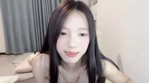 Media: Video of a young Asian woman with fair skin, long black hair, and light makeup, lying on her stomach in a room with grey curtains and white walls.