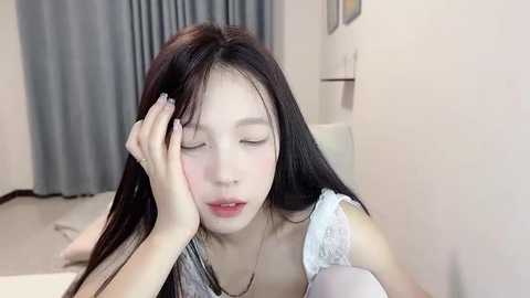 Media: A video of a young Asian woman with long black hair, fair skin, and a slender build, lying on a bed in a bedroom with beige walls and gray curtains, wearing a white lace top.