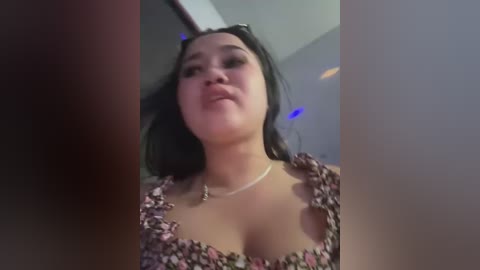 Media: Video of a young, light-skinned woman with long black hair, wearing a low-cut floral top, captured from a low angle, indoors with dim lighting.