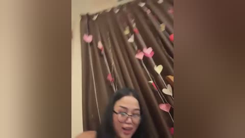 Media: Video of a woman with long black hair, wearing glasses, standing in a bathroom with brown shower curtain adorned with pink and white heart decals. She has a playful expression.