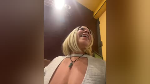 Media: Video of a blonde woman with shoulder-length hair, wearing a low-cut white top, laughing joyfully, against a yellow wall and wooden door.