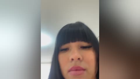 Media: Video of a young woman with straight black hair, blunt bangs, and light skin, wearing dark lipstick. The background is blurred, showing a white wall and a window with soft, diffused light.