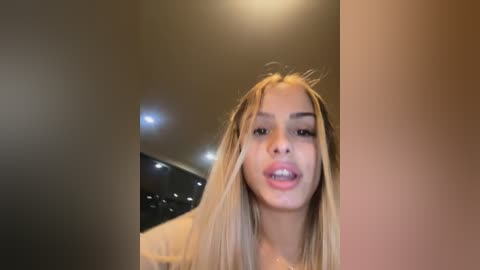 Media: A video captures a young woman with long, straight blonde hair and light skin, slightly sticking her tongue out, indoors under warm lighting.